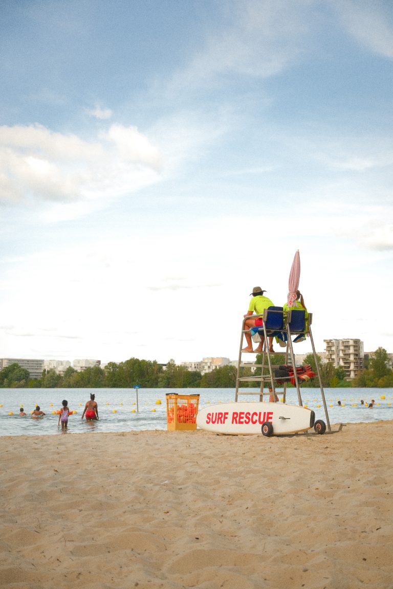 A lifeguard sits on a tall chair overlooking a sandy beach, with a ‘Surf Rescue’ sign and a rescue board nearby. Swimmers enjoy the water in the background, while beyond the lake, trees and apartment buildings complete the scene under a clear, blue sky.