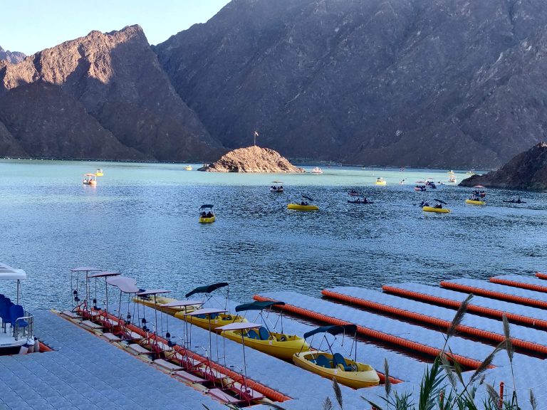 A scenic view of a lake surrounded by rocky mountains, with several pedal boats and kayaks in the water. In the foreground, a dock with more pedal boats is visible, and the background shows more boats scattered across the lake under a clear sky. Hatta Dam – Dubai