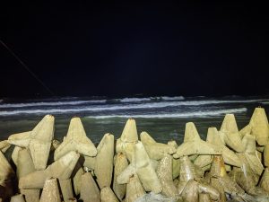 A nighttime beach scene with concrete tetrapods in the foreground and waves in the background, under a dark sky.