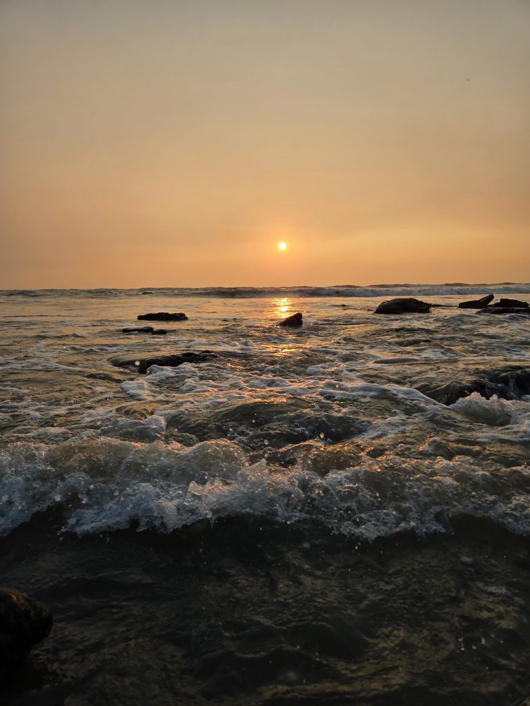 A serene view of the ocean at sunset with gentle waves crashing against rocks in the foreground. The sun is low on the horizon, casting a warm, golden glow across the water.