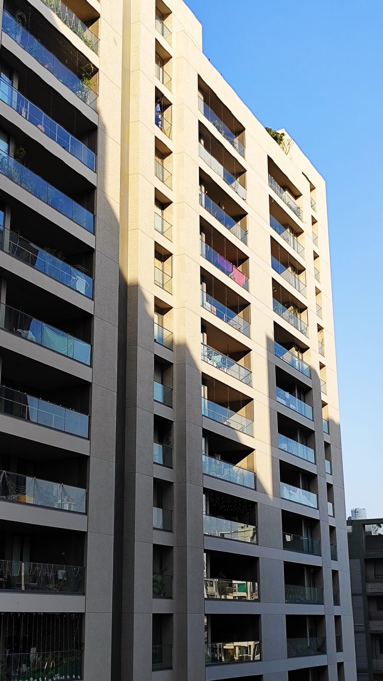 A modern high-rise residential building with glass balconies, shadowed by an adjacent structure, under a clear blue sky.