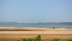 A tranquil beach scene with sandy shores leading to calm blue water. In the distance, a small boat floats on the water. A person wearing a hat walks along the water's edge, and there are distant buildings dotting the green landscape under a clear blue sky.