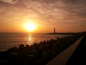 A serene sunset over the ocean with an orange sky, casting reflections on the water. Large concrete tetrapods line the shore in the foreground, and a lighthouse is visible on the distant coastline.