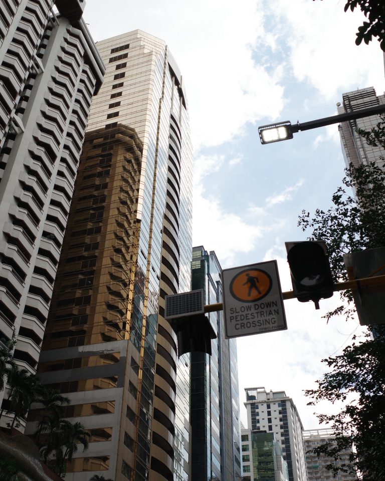 A “Slow Down, Pedestrian Crossing” sign hangs on a traffic light, with tall buildings in the background.