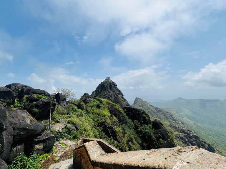 A stone pathway leading toward a mountain peak with a temple on top, with the surrounding rocky and green landscape stretching into the distance under a bright sky.