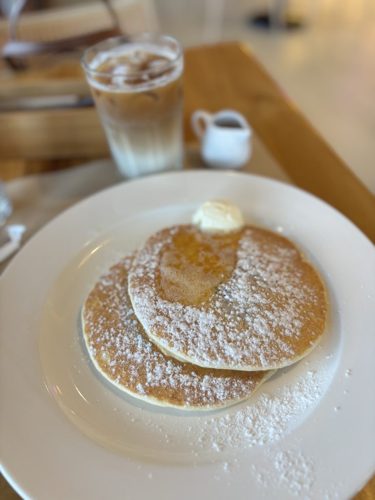 Two pancakes dusted with powdered sugar are on a white plate, topped with a small scoop of butter. A glass of iced coffee and a small pitcher of syrup are in the background on a wooden table.