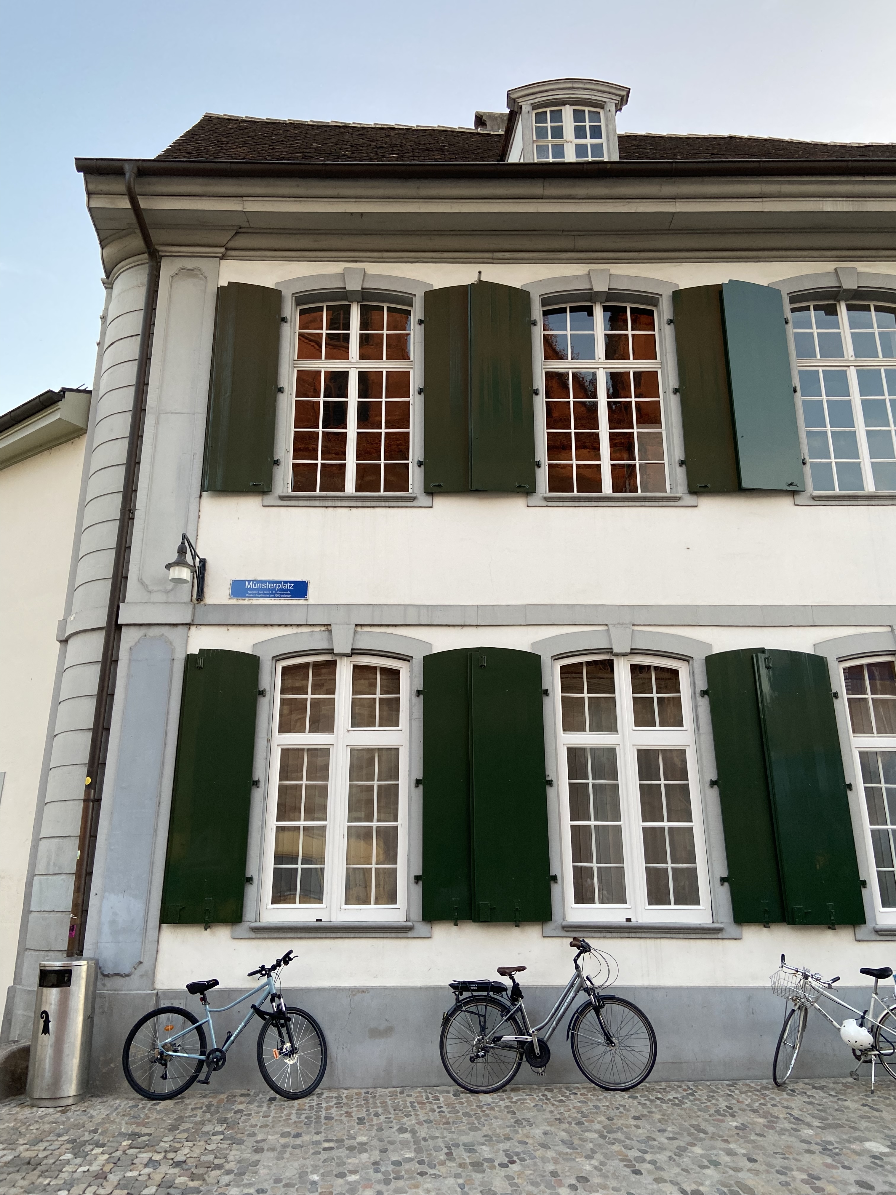 A two-story building with a sloped roof and several tall windows, some of which have green shutters. Three bicycles are parked along the cobblestone pavement in front of the building.