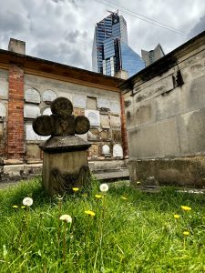 An old cemetery with tombstones, surrounded by overgrown grass and yellow dandelions in the foreground. In the background, a modern glass skyscraper towers over the scene, contrasting with the historic cemetery. The sky is cloudy. Bogotá Central Cemetery, with the Atrio building in the background.