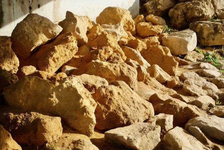 A pile of large, irregularly-shaped rocks in sunlight, with shadows casting on them. The rocks vary in size and texture, and some patches of grass are visible among them.