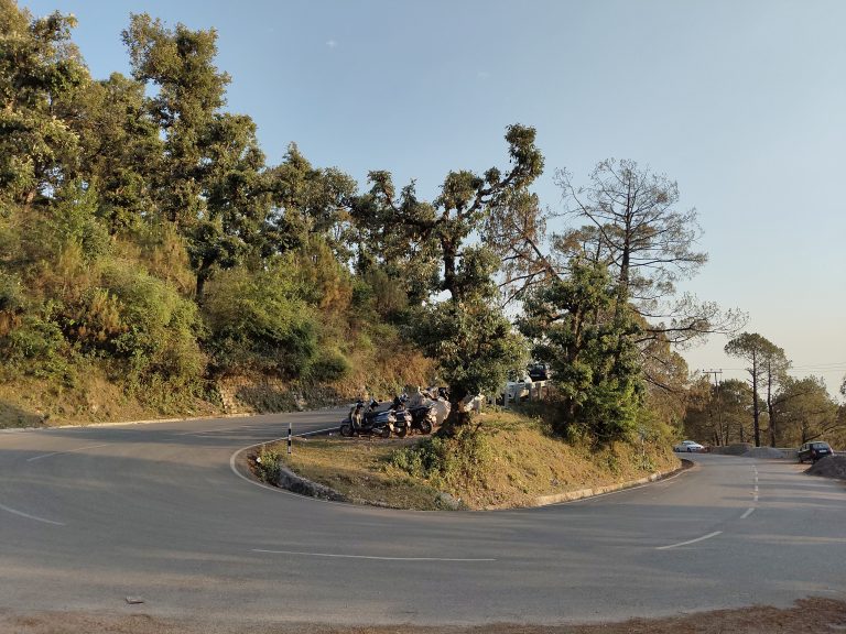 A winding road in a hilly area surrounded by lush green trees and vegetation. There are several parked motorcycles and cars at the curve of the road. The sky is clear on a sunny day.