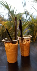 Two plastic cups filled with a frothy iced coffee beverage, topped with chocolate sprinkles, each with a black straw, set on a dark wooden table with green plants in the background.