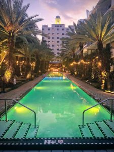 View of blue green pool with palm trees lining the left and right sides and a building in the background at Mareva1939 within National Hotel (Miami Beach, Florida)