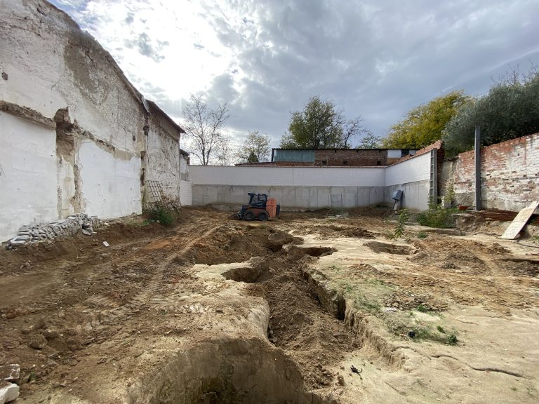 A construction site with exposed earth and tire tracks, surrounded by partially demolished and worn brick walls.