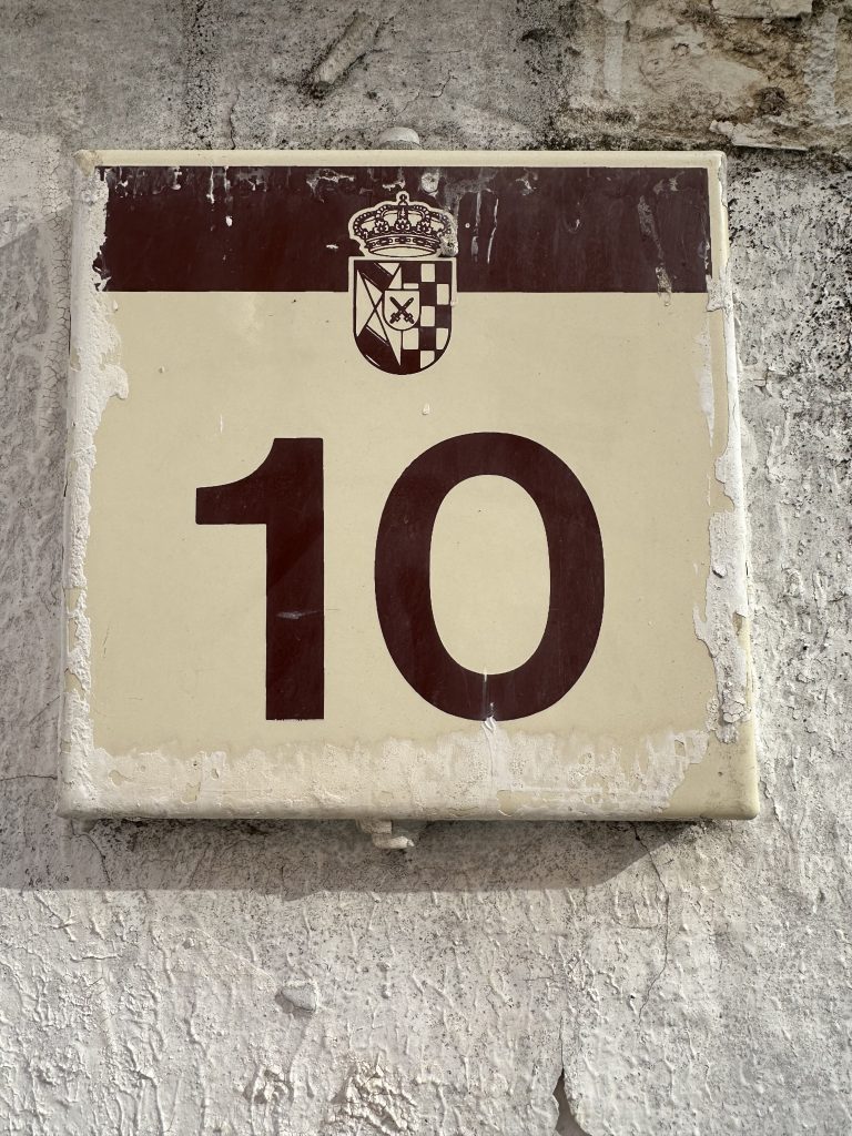 A weathered rectangular sign with the number “10” depicted in bold, dark font. Above the number is a decorative emblem featuring a crown and shield, attached to a textured wall.