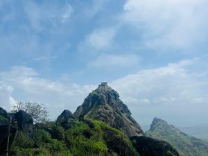 A small temple sits atop a rugged mountain peak surrounded by lush greenery, under a vast, partly cloudy blue sky.