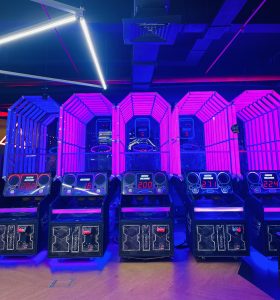 A row of arcade basketball game machines with vivid purple and blue lighting. Each machine has a digital display showing various scores, with neon signs and metal mesh cages surrounding the hoops.