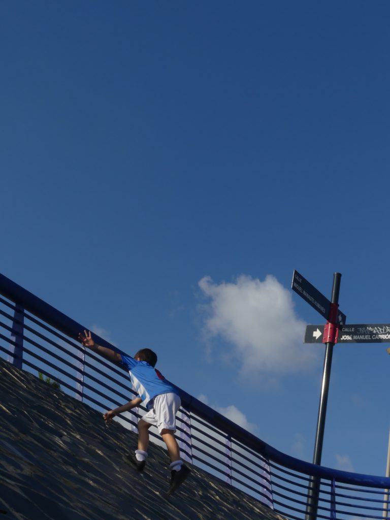 A child in a blue shirt and white shorts climbs a sloped wall, reaching towards the railing above.