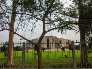 The Bangladesh parlament building stands behind a metal fence. The view is partially obscured by trees and leaves, with a grassy area in the foreground.