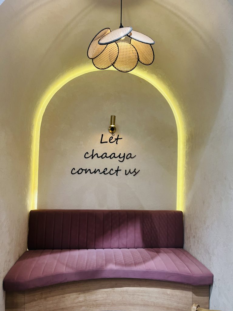 A cozy seating nook with a curved pink cushioned bench against a textured light-colored wall. Above the bench, there is a decorative pendant light shaped like overlapping petals, and the words “Let chaaya connect us” are written on the wall.