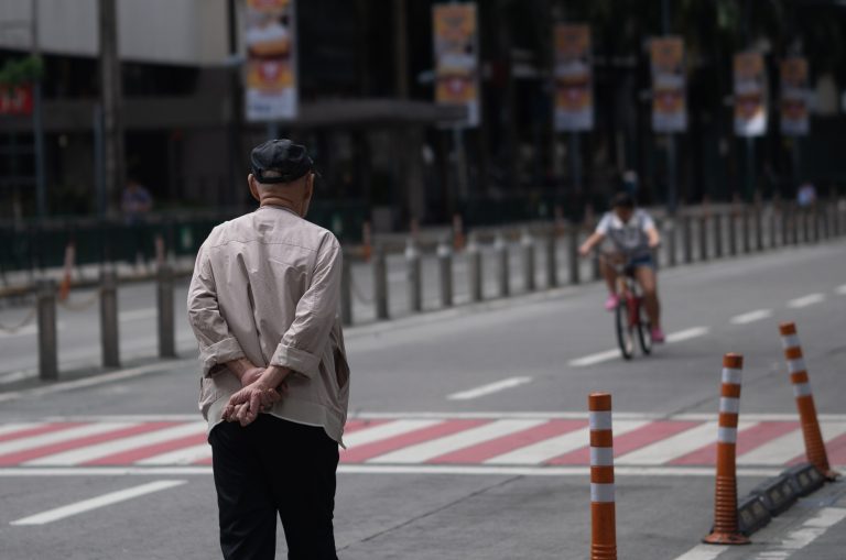 An old man walks toward a young woman on a bicycle in the distance, capturing a quiet moment that highlights their age gap