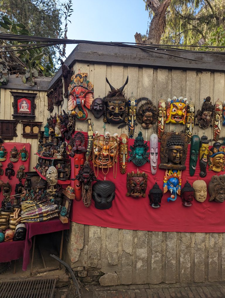 A variety of colorful masks and sculptures displayed on a wall draped with red fabric. The collection includes intricate designs featuring human faces, mythical figures, and animal motifs.