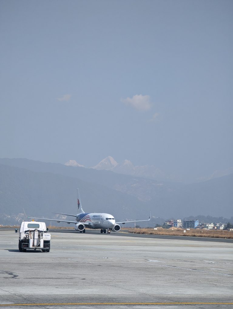 An airplane taxis on the runway of Kathmandu airport, Nepal. A small maintenance vehicle in the foreground. In the background, there are distant mountain ranges and a clear blue sky.