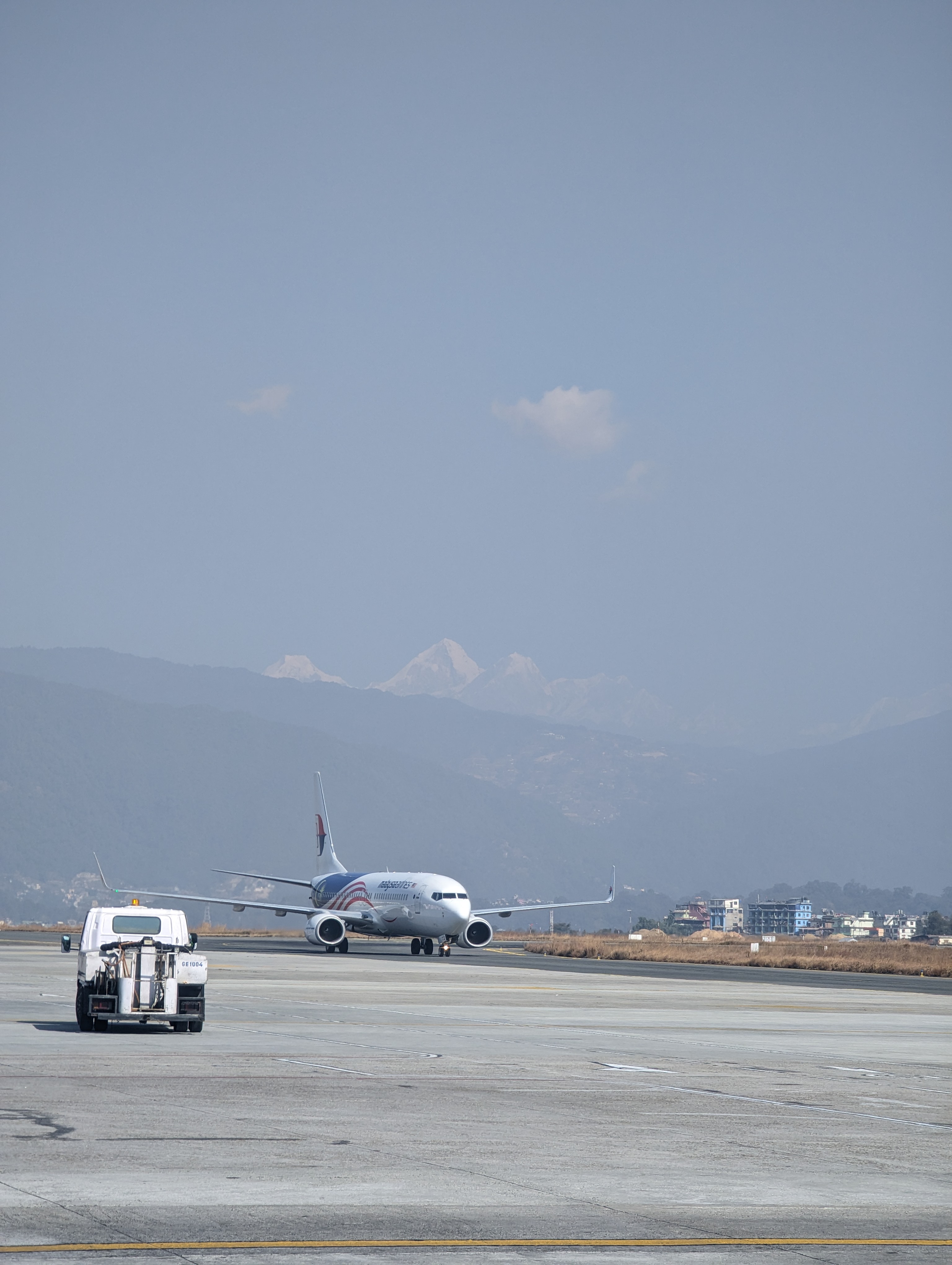 An airplane taxis on the runway of Kathmandu airport, Nepal. A small maintenance vehicle in the foreground. In the background, there are distant mountain ranges and a clear blue sky.
