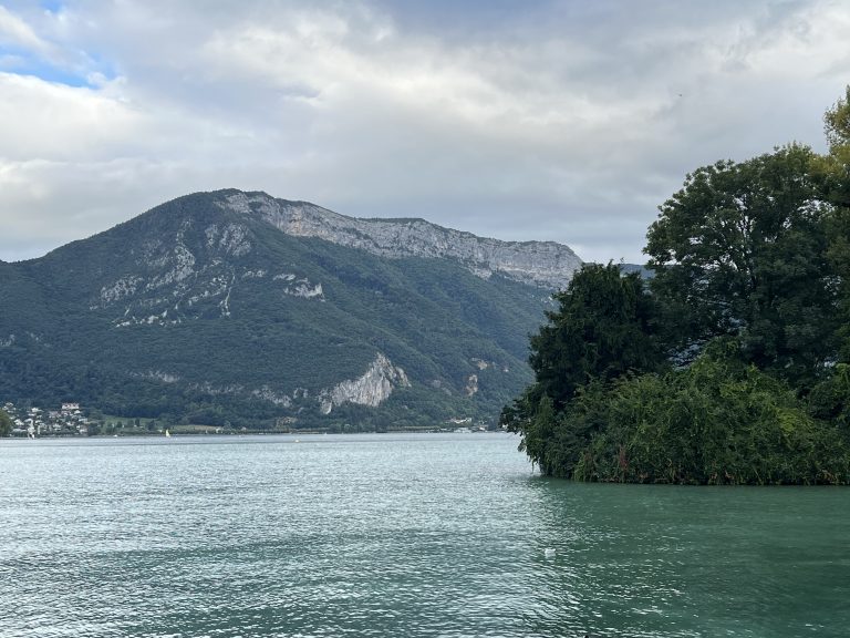 A serene landscape featuring a calm lake with greenish-blue water in the foreground, surrounded by lush greenery on one side.