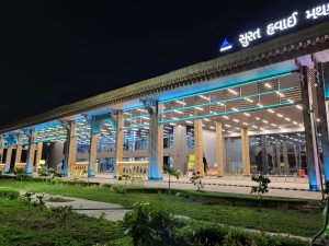 A sleek airport entrance with soft, sky-blue lighting highlights the modern design of Surat Airport. The entry gate is framed by a lush garden in the foreground, adding a touch of greenery. The combination of soft lights and natural surroundings creates a tranquil, inviting atmosphere for visitors.