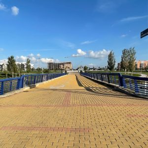 A pedestrian bridge with blue railings stretches under a clear blue sky. The path is made of yellow bricks, and trees line the sides. In the distance, there are buildings and construction cranes.