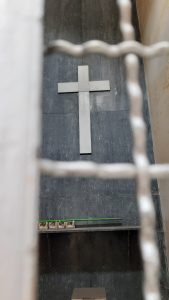 The image shows a view through a metal gate at the Central Cemetery in Bogotá. In the center, there is a large white cross mounted on a dark wall, creating a striking contrast and a solemn atmosphere. At the bottom of the image, there is a shelf with candles. The gate in the foreground adds a sense of separation and respect towards the sacred space