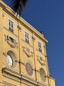 A section of an elegant yellow building with classic French architectural details, including circular and rectangular windows, decorative wreath embellishments, and ornate moldings.