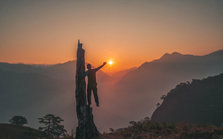 Silhouette of a man reaching toward the rising sun atop Mt. Kabunian, framed by an orange-hued sky and layered mountain ranges in the background.