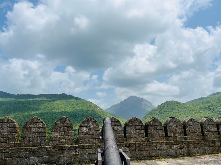 View of a historic stone wall with arched battlements and an old cannon pointing outward, overlooking lush green hills under a partly cloudy blue sky.