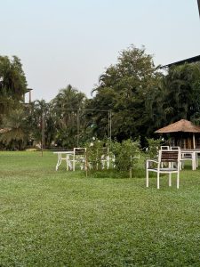 A serene outdoor setting featuring green grass and several white garden chairs and tables. The background includes lush greenery and large trees, with a clear sky overhead.