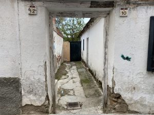 A narrow alleyway between two weathered white buildings, featuring cracked and peeling plaster. The entrance displays the numbers 12 and 10 above.