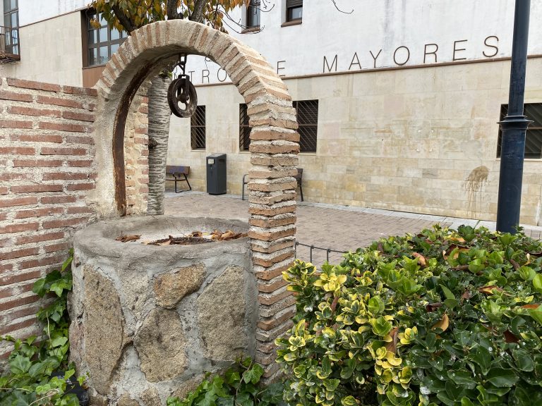 A rustic brick and stone well with a metal pulley under an arch, surrounded by green and yellow foliage.