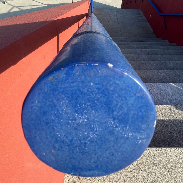 Staircase viewed from top to bottom, with a blue handrail on the left showing marks of wear and tear from the passage of time. The rustic-looking steps reflect continued use, while the perspective accentuates the depth and direction of the descent.