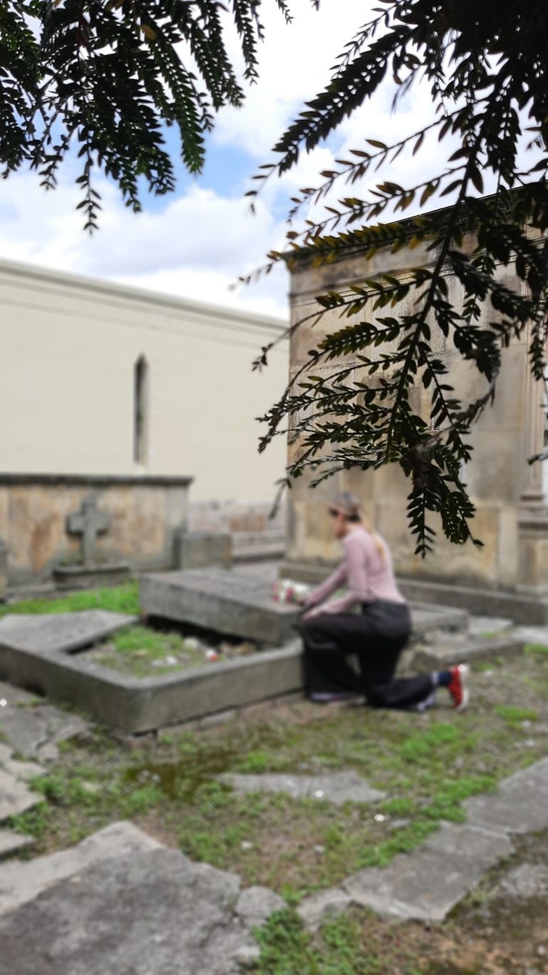 A person kneels at a gravesite in the central cemetery on Bogotá, placing flowers on a tombstone. Overhanging branches with leaves partially frame the scene. The setting is outdoors, with a stone building visible in the background.