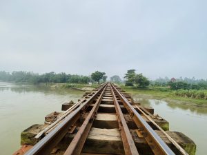 A railway track extends into the distance over a river, bordered by greenery and trees, under a cloudy sky.