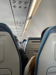 Interior view of an airplane cabin, showing rows of blue and grey seats. Passengers are seated and viewed from behind, with overhead compartments and air vents visible above.