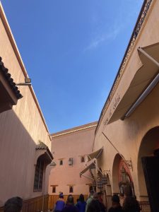 A narrow alleyway with peach-colored walls and an archway at Marrakesh medina featuring decorative lanterns and awnings. A group of people is walking through the passage under a clear blue sky.