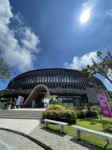 A modern, circular building with a stone facade and large, vertical windows under a bright blue sky. The sun is shining, creating a sunburst effect in the upper right corner. In front of the building are stairs leading up to an entrance. 