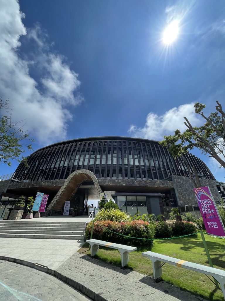 A modern, circular building with a stone facade and large, vertical windows under a bright blue sky. The sun is shining, creating a sunburst effect in the upper right corner. In front of the building are stairs leading up to an entrance.