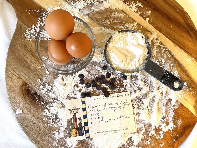 A wooden cutting board with three brown eggs in a glass bowl, a black measuring cup filled with flour, scattered chocolate chips, and a handwritten recipe card for chocolate chip cookie dough pie. Flour is sprinkled across the surface.