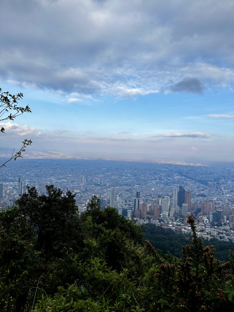 Aerial view of Bogota city skyline with a mix of modern high-rise buildings surrounded by lush trees in the foreground. The sky above is partly cloudy, with patches of blue visible.