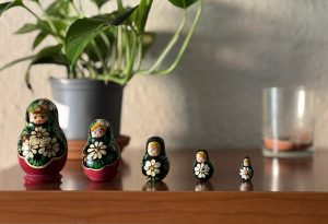 A set of matryoshka dolls in the foreground, on a wooden piece of furniture. In the background, blurred, there is a potted plant and a candle in a glass, almost spent.