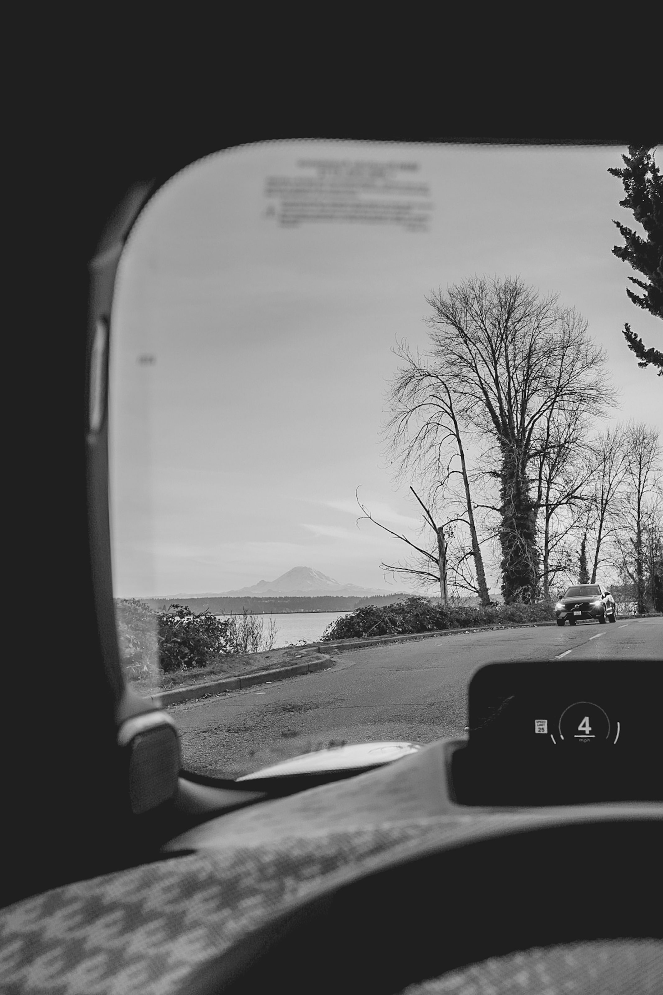 Black and white photo of a view of Mount Rainier appearing in the distance from inside a car driving alongside a two lane road with another car approaching.