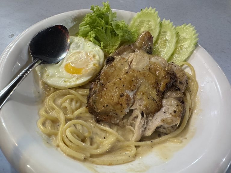 A plate of creamy spaghetti carbonara with grilled chicken on top, accompanied by a fried egg. The dish is garnished with lettuce and slices of cucumber, and a spoon rests on the plate.