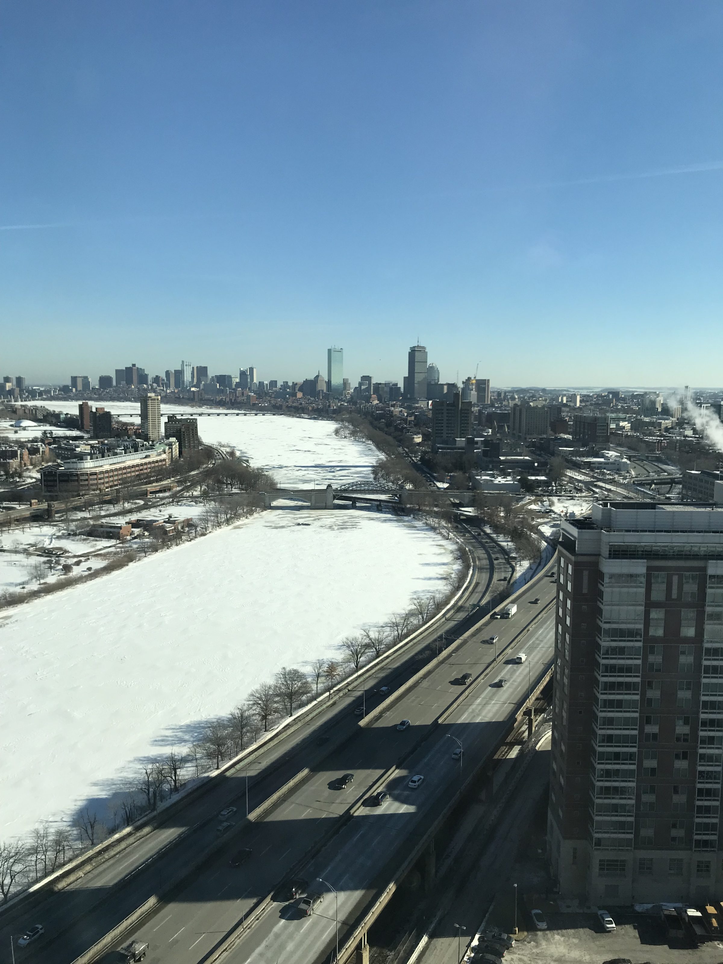 A frozen, snowy Charles River seen from above with the downtown Boston skyline in the distance.